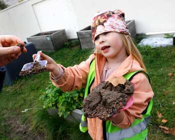Дитина у світловідбиваючій окулярах тримає маленьку пластикову іграшкову лопатку з частиною глиняного коржика на ній. Решту глиняного коржика вона тримає в іншій руці.